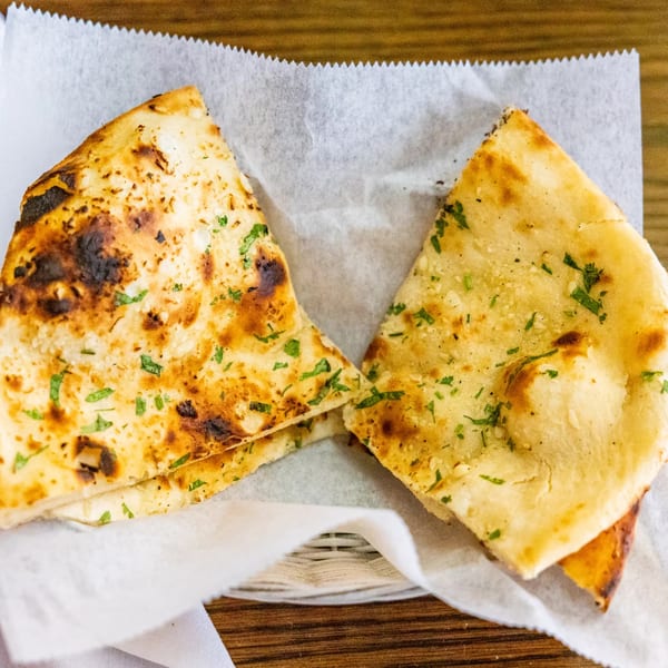 Two pieces of golden-brown Indian naan bread, garnished with green herbs, served in a white paper-lined basket.