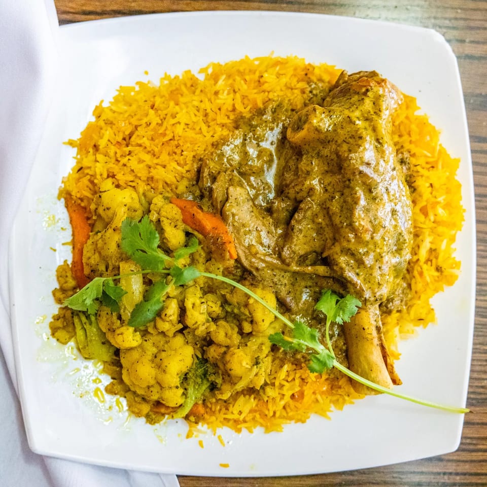 Indian lamb shank with green curry, yellow rice, and mixed vegetables on a white plate.