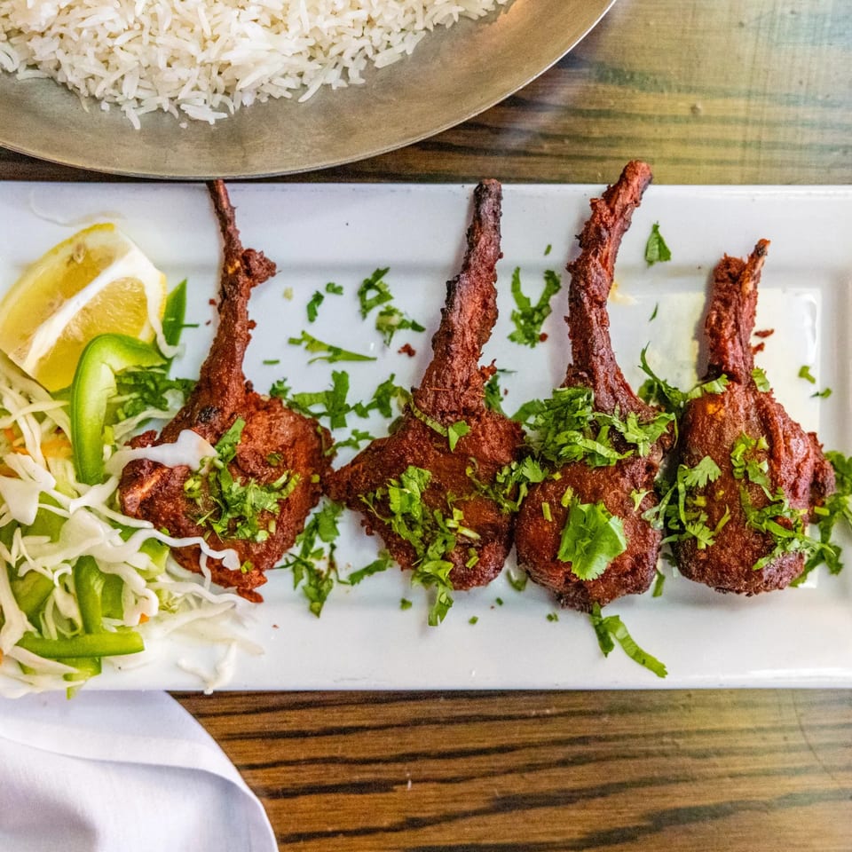 Four Rasoi Indian lamb chops, garnished with cilantro, lemon, and shredded cabbage, alongside rice.
